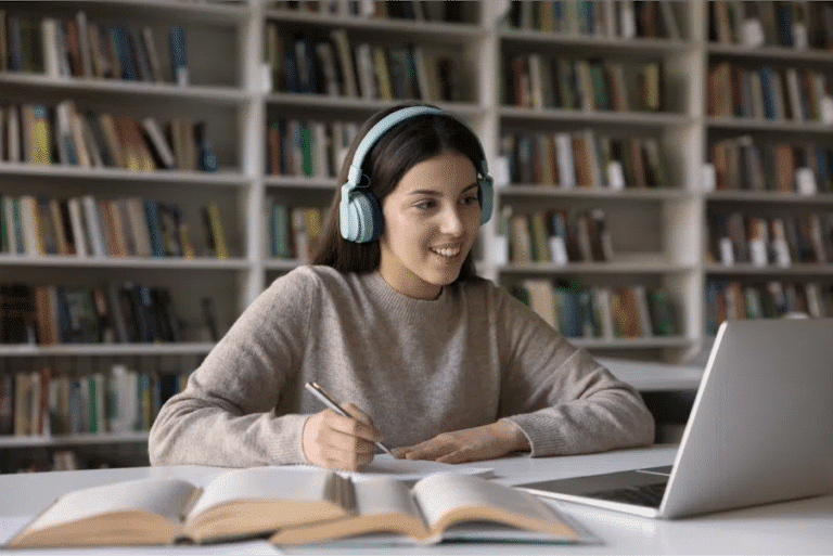 A woman wearing headphones is sitting at a desk with a laptop in front of bookshelves.